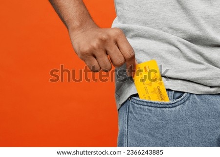 Croped close up shot photo of young man he wearing t-shirt casual clothes holding in hand put mock up of credit bank card into pocket isolated on orange red color background studio. Lifestyle concept