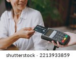 Croped young smiling fun happy woman wearing white shirt hold wireless bank payment terminal to process acquire credit card sit alone at table in coffee shop cafe restaurant indoor. Focus on machine