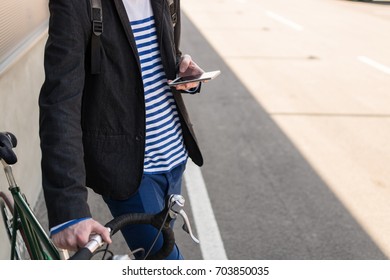 Cropped angled view image of a young attractive man wearing casual clothes holding his bicycle on the street against a wall while using smart phone. Style for a busy man on the move