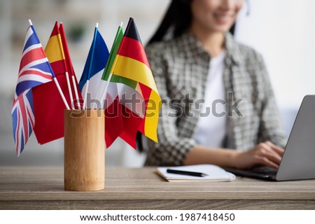 Cropped of asian young woman foreign language teacher sitting at workdesk and using laptop, selective focus on international flags. Foreign language online course, class, e-education concept