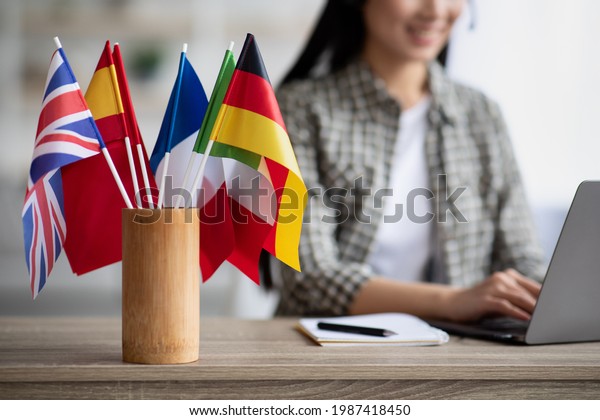 Cropped of asian young woman foreign language\
teacher sitting at workdesk and using laptop, selective focus on\
international flags. Foreign language online course, class,\
e-education concept
