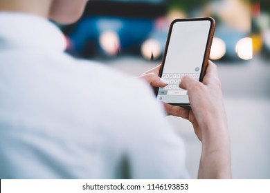 Cropped back view of young woman holding smartphone in hands and typing text on keyboard of modern device with blank display.Hipster blogger messaging with followers on telephone with blank screen