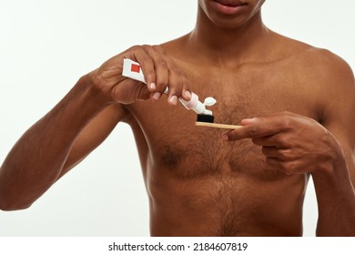 Cropped Black Guy Pouring Toothpaste On Toothbrush Before Brushing Teeth. Obscure Face Of Young Man With Naked Torso. Dental Care And Hygiene. Isolated On White Background. Studio Shoot