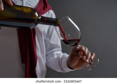 Cropped Of Black Male Winemaker Or Sommelier Pouring Red Wine In Glass From Bottle. Concept Of Wine Tasting. Viticulture And Winemaking. Isolated On Grey Background. Studio Shoot