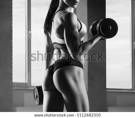 Cropped black and white photo of strong fit model training in gym , doing fitness exercises with heavy dumbbells. Having athletic muscles, healthy body and tanned skin. Looking strong, feeling good.