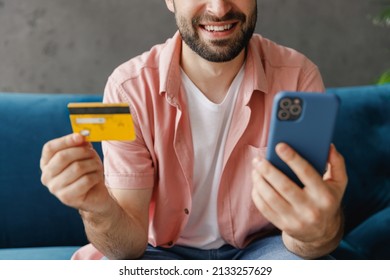 Cropped Close Up Fun Young Man In Casual Clothes Use Mobile Cell Phone Hold Credit Bank Card Shopping Online Order Delivery Book Tour Sitting On Blue Sofa At Home Flat Indoor Rest Relax On Weekends