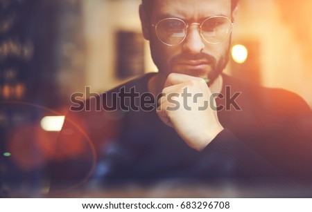 Cropped close up image of pensive bearded entrepreneur in optical spectacles for better views dressed in black sweatshirt.Pondering businessman in trendy eyeglasses thinking on blurred background