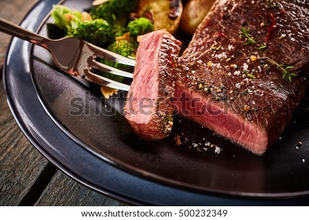 Cropped close up on cut of medium rare cooked beef steak on fork in plate with broccoli