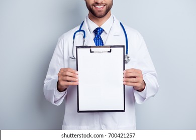 Cropped close up photo of bearded smiling intern, who is holding the clipboard with empty paper. Doc is wearing white uniform and a tie, stands on a light white background