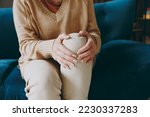 Cropped close up photo of pensioner woman wear casual clothes sits on blue sofa hold knee suffer from leg knee pain inflammation stay home flat spend free spare time in living room indoor grey wall