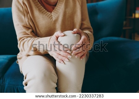 Cropped close up photo of pensioner woman wear casual clothes sits on blue sofa hold knee suffer from leg knee pain inflammation stay home flat spend free spare time in living room indoor grey wall