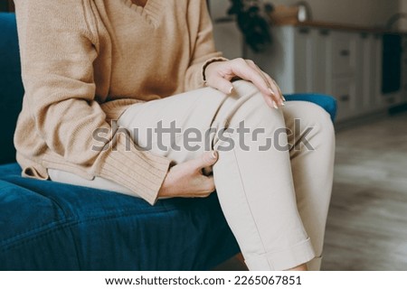 Cropped close up photo of pensioner woman wearing casual clothes sit on blue sofa hold knee suffer from leg knee pain inflammation stay home flat spend free spare time in living room indoor grey wall