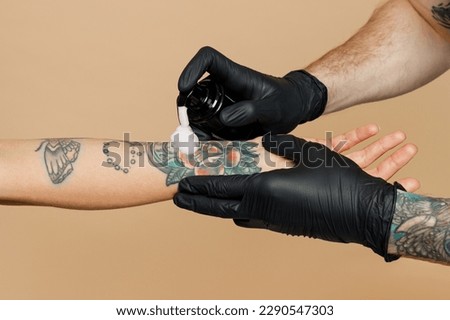 Cropped close up photo shot of tattooer master artist tattooed man wearing black sterile gloves applying cream on female hand after making colored tattoo art on body isolated on plain beige background