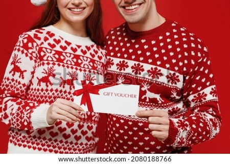 Cropped close up photo young couple friends two man woman in sweater hat hold gift certificate coupon voucher card for store isolated on plain red background. Happy New Year 2022 celebration concept