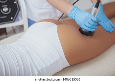 Cropped close up of professional cosmetologist using ultrasound cavitation machine on a female client. Unrecognizable woman getting contouring anti-cellulite body treatment at beauty salon