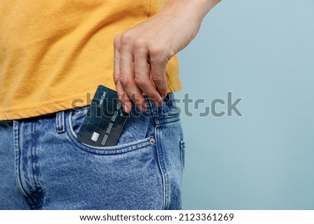 Cropped close up rich man 20s wearing yellow t-shirt hold in hand put credit bank card to jeans denim pocket isolated on plain pastel light blue background studio portrait. People lifestyle concept.