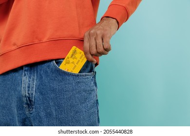 Cropped close up shot photo of cool african american man in orange shirt holding in hand put into denim jeans pocket credit bank card isolated on plain pastel light blue background studio portrait.