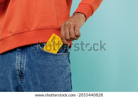 Cropped close up shot photo of cool african american man in orange shirt holding in hand put into denim jeans pocket credit bank card isolated on plain pastel light blue background studio portrait.