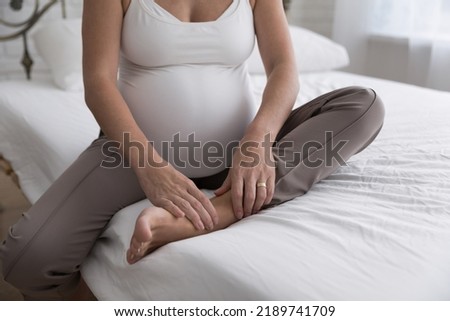 Cropped close up shot unrecognizable pregnant female sits on bed touch, massages her swollen foot or aching ankle, suffering from leg cramps, muscles pain due to overweight in late pregnancy concept