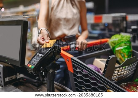 Cropped close up woman hand shopping at supermaket put credit card to wireless modern bank payment terminal process acquire payments near cashier checkout with cart inside store. Purchasing concept