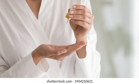 Cropped close up woman holding bottle with cosmetics moisturizing serum or oil, facial essence, using pipette, young female in bathrobe doing cosmetology beauty skincare routine procedure