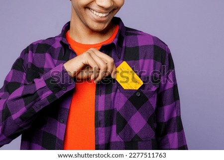 Cropped close up young man of African American ethnicity wear casual shirt orange hat hold in hand mock up of credit bank card put into pocket isolated on plain pastel light purple color background