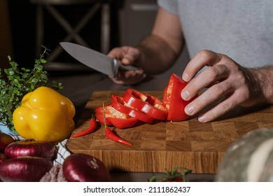 Cropped closeup portrait of male hands cutting red bell pepper in the kitchen with knife on wooden cutting board. Onion, pumpkin, hot pepper and greenery are on the table.