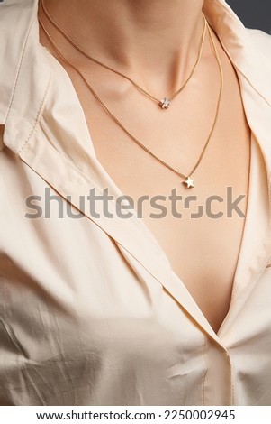 Cropped close-up portrait of a woman, demonstrating a double gold necklace with star pendants in a beige shirt. The necklace consists of two different chains. The girl is on a gray background.        