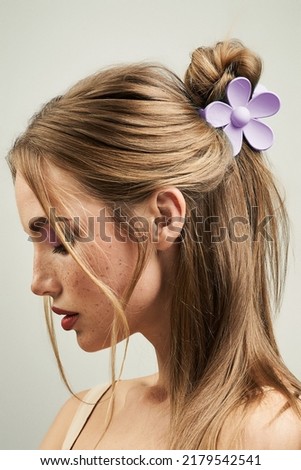 Cropped close-up shot of a blonde girl with a claw clip in her hair. Portrait of a young woman with a violet flower hair claw clip on a pastel background. Side view.          