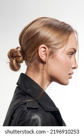 Cropped Close-up Shot Of A Blonde Young Woman With A Chignon. The Woman In The Black Jacket With The Updo Is On A White Background. Side View.