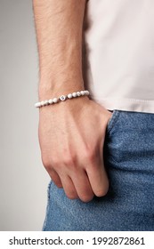 Cropped close-up shot of male wrist with beaded bracelet made of white marble stone and decorated with silver beads and charm with letter G. Man in beige t-shirt put his hand in the pocket.
