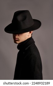 Cropped close-up shot of a man in a black wide-brimmed fedora. A young man in black clothes and a felt hat covering face is posing on a dark background. Side view.              