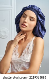 Cropped close-up shot of a woman in a dark blue sleep cap with a wide elastic band. A girl in a satin hair bonnet and in white lingerie touches her neck with fingers on a light background. Front view.