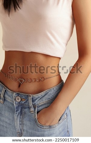 Cropped close-up shot of a woman with a silver waist chain. A girl in a white top, a belly chain with heart details and her hand in a pocket of blue jeans is on a pastel background. Front view.