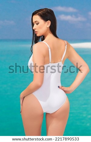 Cropped close-up shot of a young woman in a white one-piece swimsuit with a tie in front. A beautiful dark-haired girl in a cutout swimsuit is posing by the sea. Back view.                        
