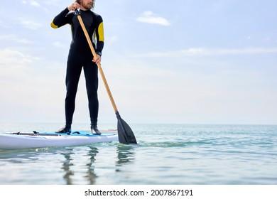 Cropped Confident Athlete man in black wetsuit doing stand-up paddleboard in ocean, engaged in sport. Athlete paddleboarding on SUP surf board. Copy space. Bearded guy using paddles - Powered by Shutterstock