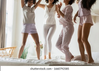 Cropped diverse girls best friends wearing pajamas jumping on bed, having fun at sleepover or hen-party, beautiful young women dancing in bedroom, celebrating, enjoying leisure time together