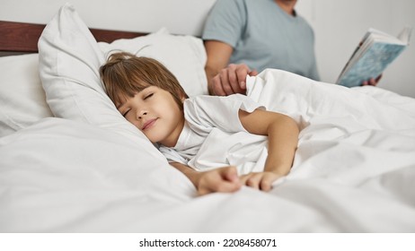 Cropped father with book covering his sleeping caucasian little son with blanket in bed at home. Family relationship and spending time together. Fatherhood and parenting. Modern domestic lifestyle - Powered by Shutterstock