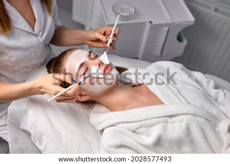 Cropped female doctor cosmetologist or dermatologist making face mask in cosmetology office. Professional Beautician applying face mask on caucasian woman face lying on bed in bathrobe. copy space