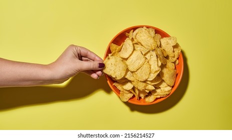 Cropped Female Hand Taking Delicious And Appetizing Potato Chip From Bowl. Unhealthy Eating And Junk Food. Crispy Snack For Leisure. Isolated On Yellow Background. Studio Shoot. Copy Space. Top View