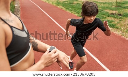 Cropped female trainer noting time on smartwatch of running caucasian boy on running track outdoors. Sportswoman with tattoos and child wearing sportswear. Modern healthy and sports lifestyle
