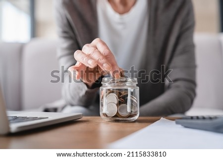 Cropped focused shot. Savings concept. Nest egg of old elderly senior woman grandmother saving money, economizing pension, mortgage loan at home using laptop and putting coin into moneybox