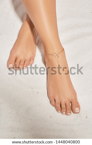 Cropped front shot of girl's legs with french pedicure, wearing golden ankle bracelet, decorated with golden insertion in view of tied bow. The lady is crossing her legs, lying on the sandy platform.