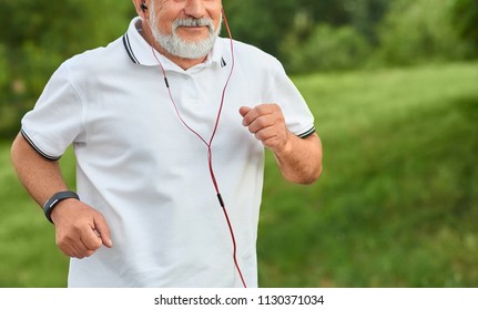 Cropped frontview of running old man having white beard. Wearing classic white shirt with dark blue stripes, sport watch and red headphones. Looking sporty, fit, healthy. Green, grassy city park.