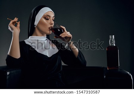 Cropped half-turn shot of a nun, sitting on a chair. She's wearing dark nun's clothing. The nun is drinking wine and holding cigarette in her hand. There is a bottle with alcohol on a chair armrest. 