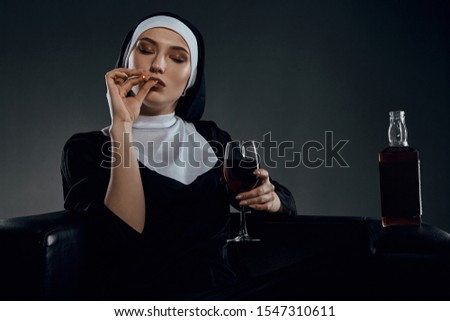 Cropped half-turn shot of a nun, sitting on a black chair. She's wearing dark nun's clothing. The nun is smoking and holding glass of wine in her hand. There is a bottle with alcohol on chair armrest.