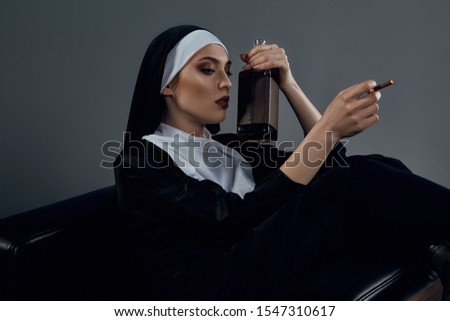 Cropped half-turn shot of a nun, sitting on a black chair. She's wearing dark nun's clothing. The nun is smoking and holding bottle of whiskey in left hand. 