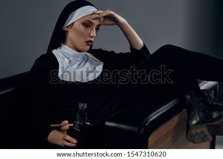 Cropped half-turn shot of a nun, sitting on a black chair. She's wearing dark nun's clothing. The nun is holding bottle of whiskey and cigarette in left hand. Her left hand is on a  forehead. 