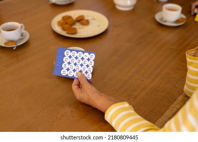 Cropped Hand Of Biracial Senior Woman Holding Bingo Card At Dining Table In Nursing Home. Cookie, Coffee, Luck, Leisure Game, Unaltered, Support, Assisted Living And Retirement Concept.