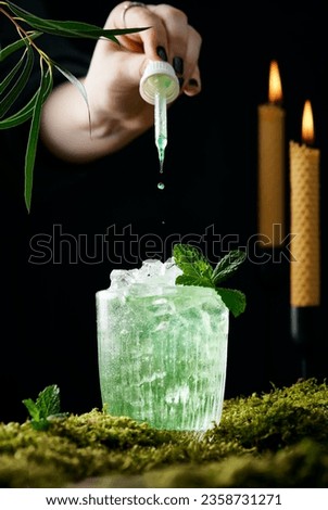Cropped hand dripping drops into very icy light green cocktail. Cocktail standing on the moss, black background, lit burning candles. Vertical lifestyle images with a mysterious atmosphere.